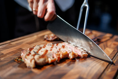 Persoon snijdt rosé gegrilde steak met groot koksmes op kopshouten snijplank in buitenlucht