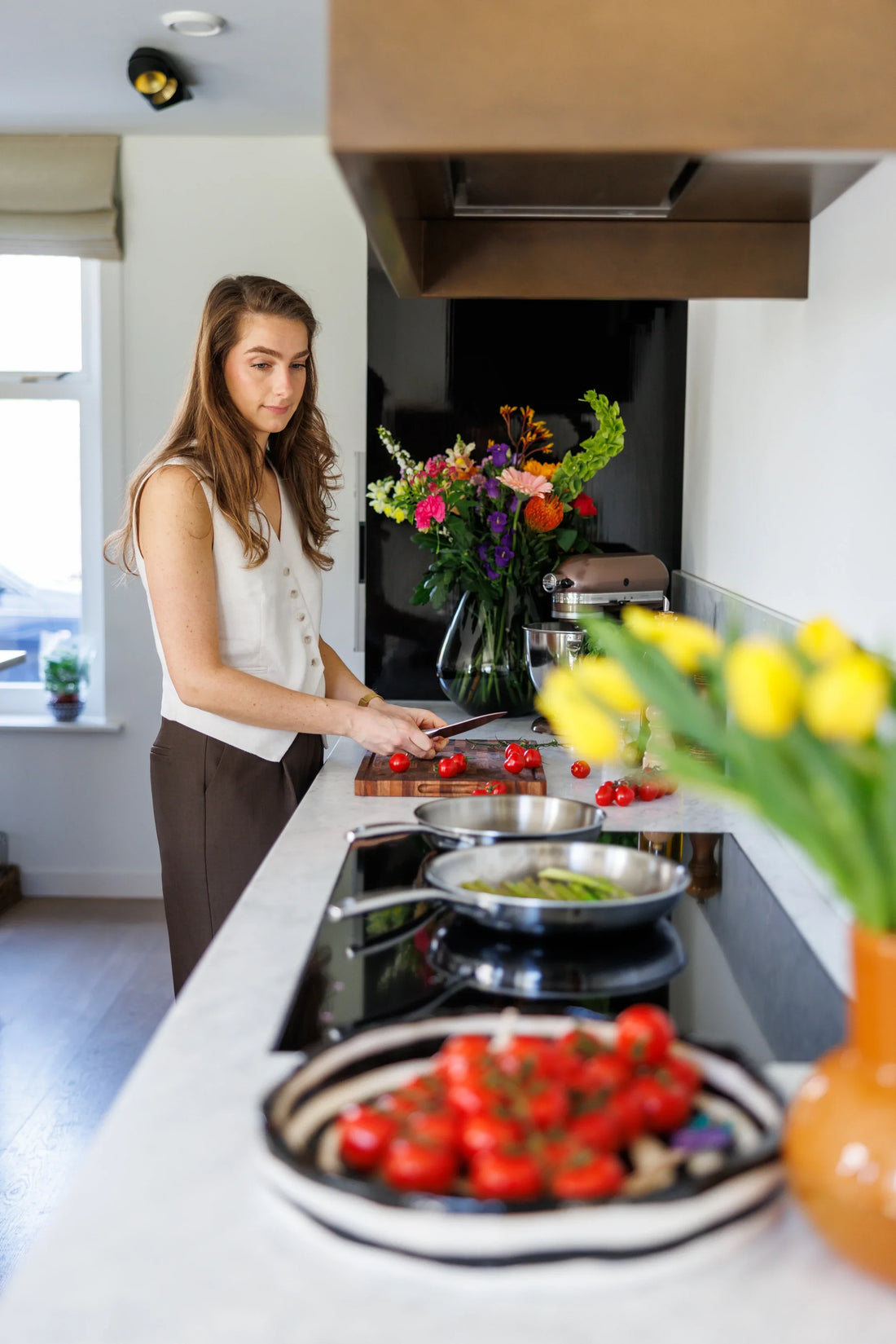 Vrouw snijdt tomaten op houten snijplank naast twee RVS koekenpannen op inductie in lichte keuken