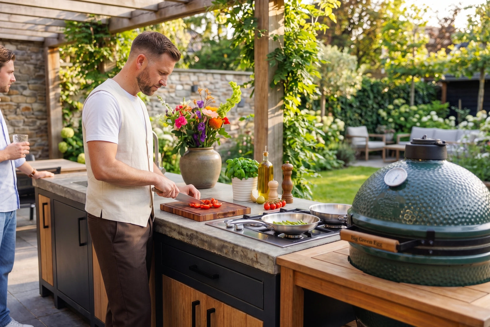 Man snijdt tomaten op een houten snijplank in een buitenkeuken, met RVS koekenpan en kamado-barbecue