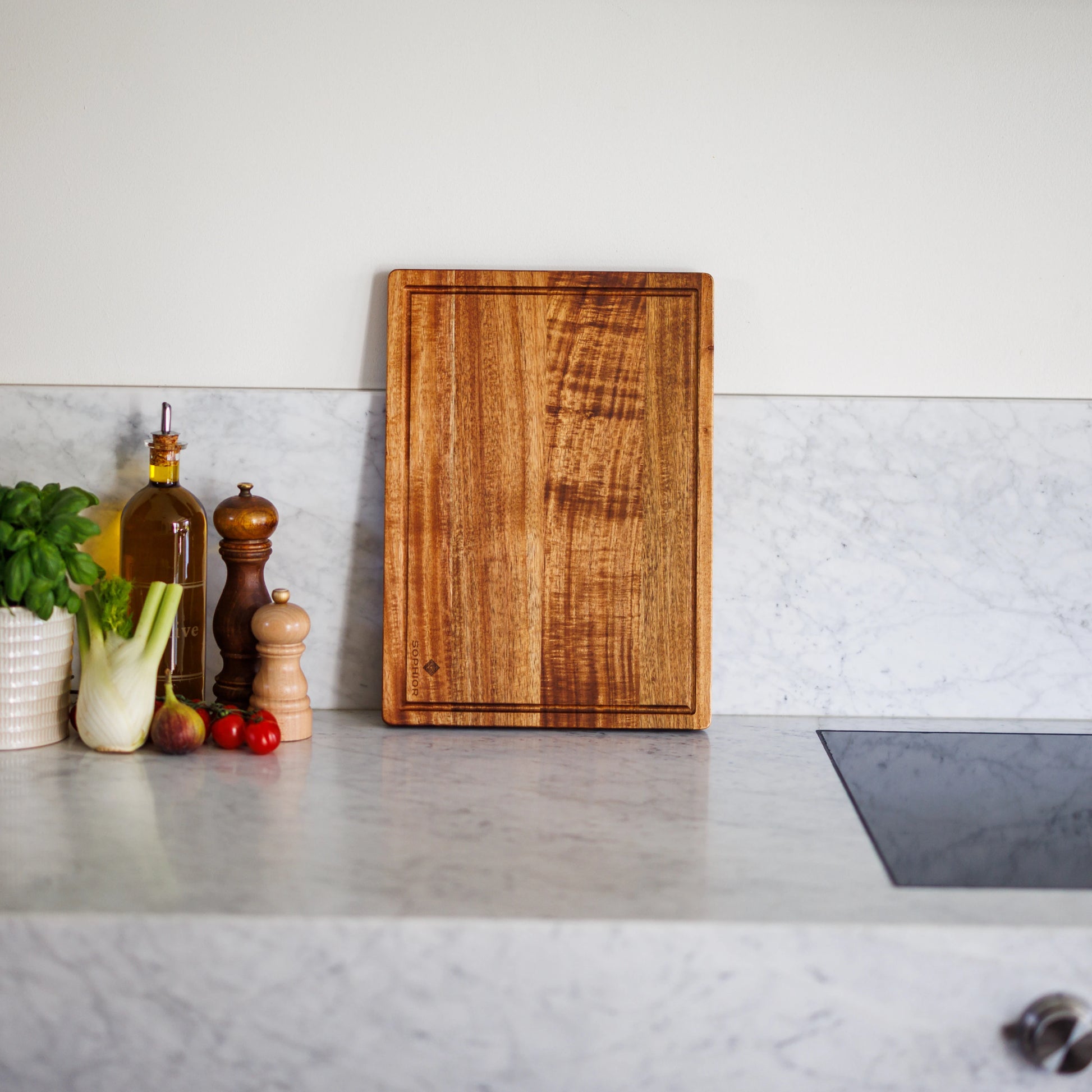 Houten Snijplank Acacia – 42×30×2,5 cm rechtop op marmeren werkblad met basilicum, olijfolie en venkel