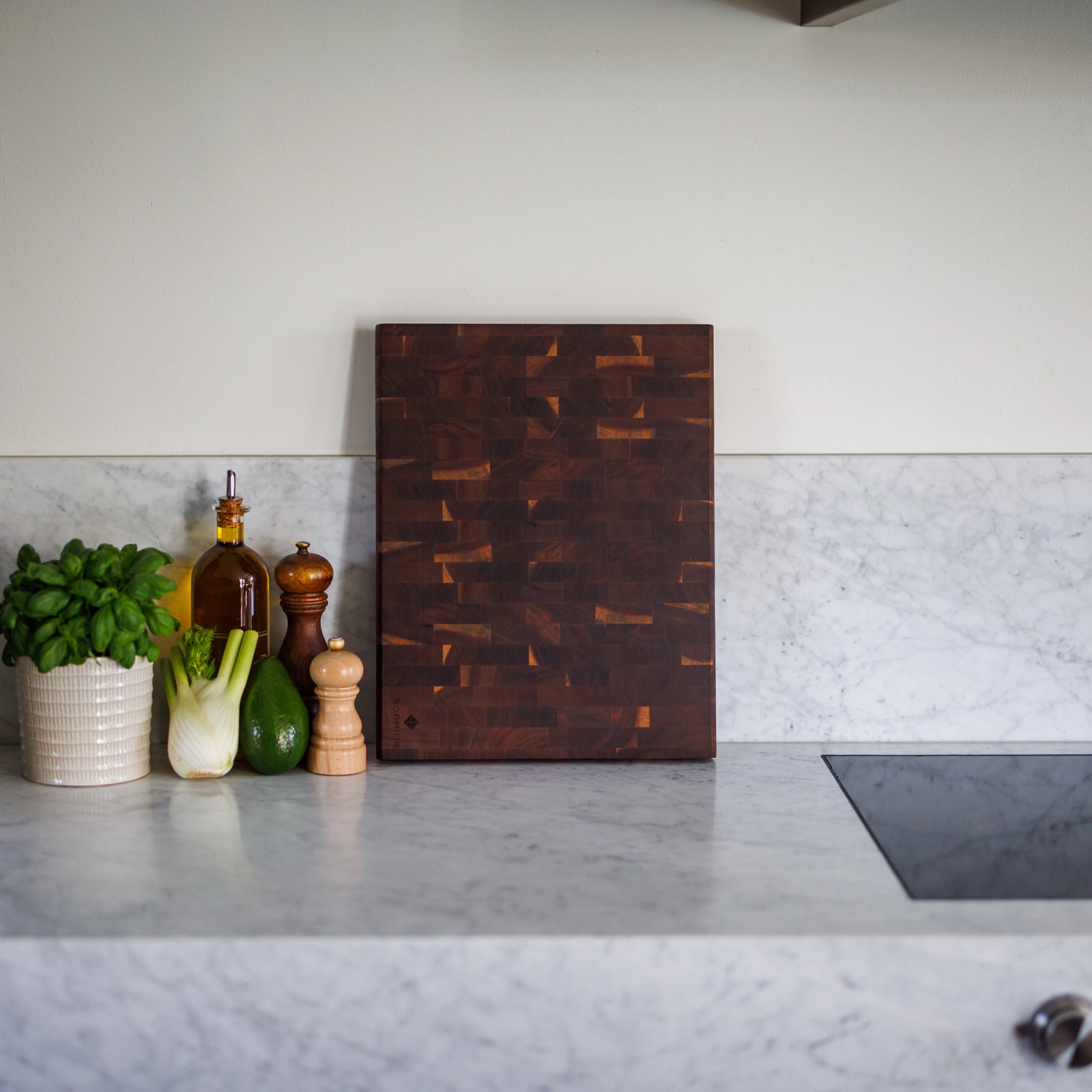 Houten Snijplank Acacia Kopshout – 45×35×4 cm rechtop op marmeren werkblad met basilicum, olijfolie en peper- en zoutmolens